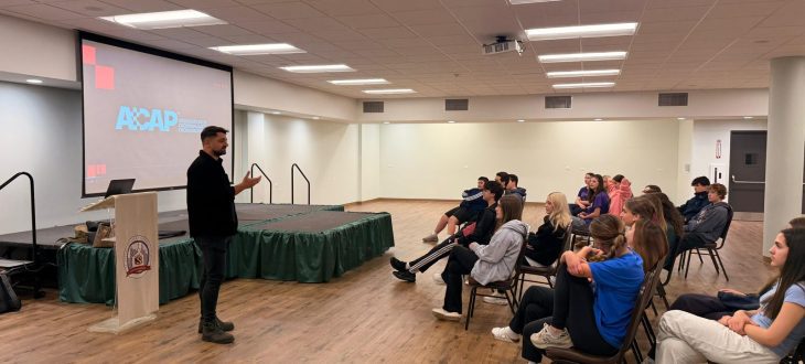 A guest speaker stands beside a podium and presents to Klub Stepinac teens seated on chairs in the parish hall, with the ACAP (Association of Croatian American Professionals) slide on the screen behind him.