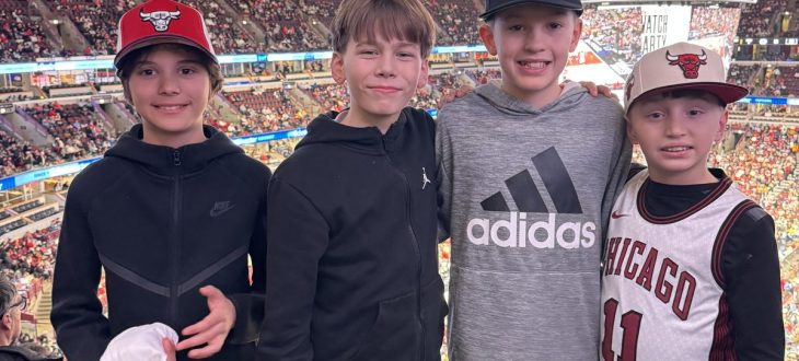 Four boys in Bulls caps and jerseys pose at a railing high in the United Center, with the Bulls' championship banners hanging above the court.