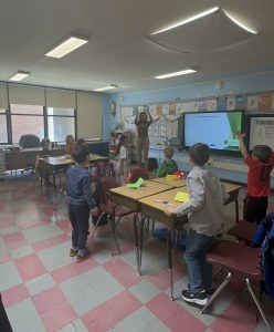 A workshop leader leads younger students in an action game at the front of a classroom, a presentation on the screen beside her.