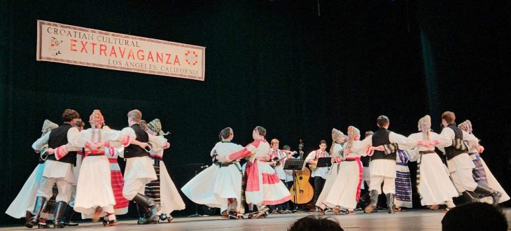 (English) Dancers in Croatian folk costumes and beaded headpieces dancing a kolo on stage beneath the "Croatian Cultural Extravaganza, Los Angeles, California" banner, tamburaši playing behind them.