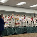The youngest students in Croatian folk costumes stand on a small stage holding hand-drawn posters of hearts, flowers and rainbows, with letters spelling MAMA, while a teacher stands beside the stage with the program in hand.