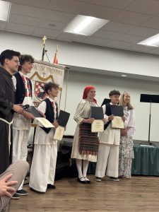 A friar, a teacher and four students in Croatian folk costumes hold diplomas in front of the St. Jerome parish banner.